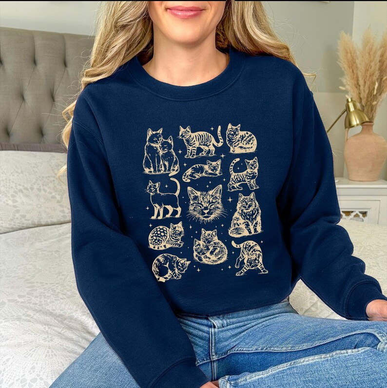 May include: Navy blue sweatshirt featuring a beige illustration of various cat poses. The design includes cats sitting, standing, and lying down. The sweatshirt is made of a soft material, perfect for casual wear.
