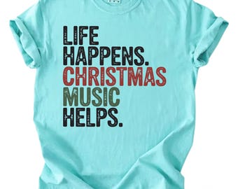 May include: A light blue t-shirt with the text "LIFE HAPPENS. CHRISTMAS MUSIC HELPS." in black, red, and green lettering. The shirt has a crew neck and short sleeves, and the fabric is slightly wrinkled.