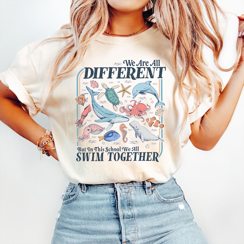 May include: Cream-colored t-shirt with a colorful graphic featuring various sea creatures like whales, dolphins, and octopuses. The text reads "We Are All Different But In This School We All Swim Together."