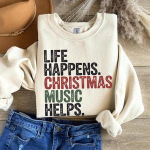 May include: Cream-colored sweatshirt with the text "LIFE HAPPENS. CHRISTMAS MUSIC HELPS." in black, red, and green lettering. The sweatshirt is paired with blue denim jeans and a brown hat.