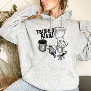 May include: A light grey hoodie with the text "Trashed Panda" and a graphic of a raccoon, a toilet, a bin, and a spilled bottle. The hoodie is made of a soft material.