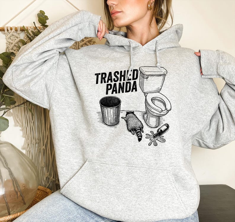 May include: A light grey hoodie with the text "Trashed Panda" and a graphic of a raccoon, a toilet, a bin, and a spilled bottle. The hoodie is made of a soft material.