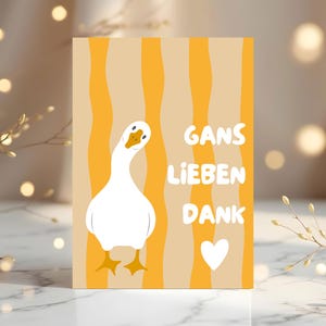 May include: A rectangular greeting card featuring a white goose with an orange beak and feet, set against a background of vertical wavy orange and beige stripes. The text "GANS LIEBEN DANK" is printed in white, accompanied by a white heart icon.
