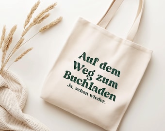 May include: A cream-coloured canvas tote bag with two shoulder straps, featuring dark green text that reads "Auf dem Weg zum Buchladen Ja, schon wieder." The bag is made of a woven fabric and has a simple, rectangular shape with a flat base.