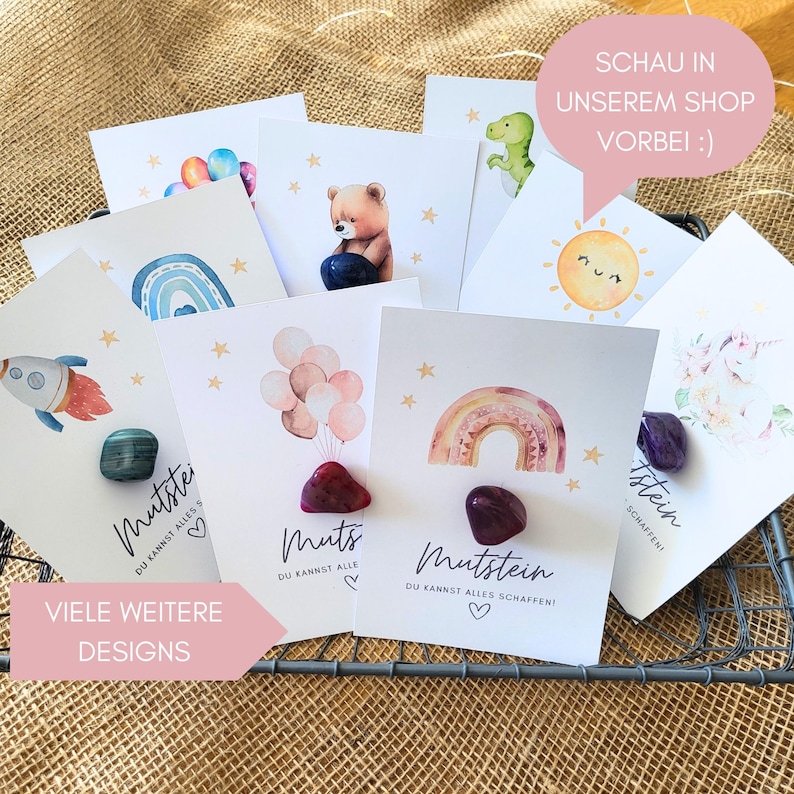 May include: A collection of white rectangular cards featuring watercolor illustrations of rainbows, balloons, a rocket, a bear, a sun, and a unicorn. Each card displays the German text Mutstein and a polished gemstone attached to the center.