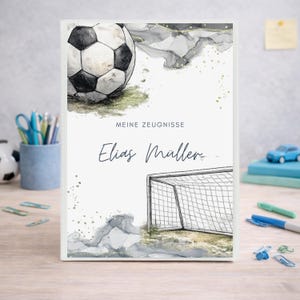 Könnte beinhalten: Ein personalisiertes Notizbuch mit Fußballmotiv, das einen Fußball und ein Torpfosten zeigt. Auf dem Cover stehen die Worte "MEINE ZEUGNISSE" und der Name "Elias Müller". Das Design beinhaltet Aquarell-Akzente in Grau und Gold.