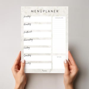 Könnte beinhalten: Ein weißer Wochen-Menüplaner mit dem Titel "MENÜPLANER". Er bietet Abschnitte für jeden Wochentag, eine Einkaufsliste und Notizen. Der Planer wird von zwei Händen gehalten.