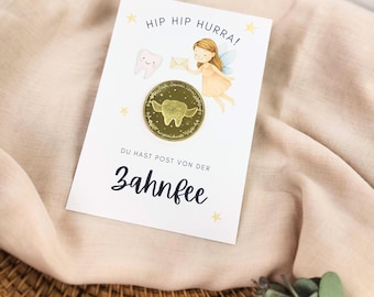 Op de afbeelding: Een witte kaart met de tekst "HIP HIP HURRA!" en "DU HAST POST VON DER ZAHNFEE" is versierd met een tand, een fee en een gouden munt. De munt heeft een tand met vleugels. De kaart ligt op een beige doek.