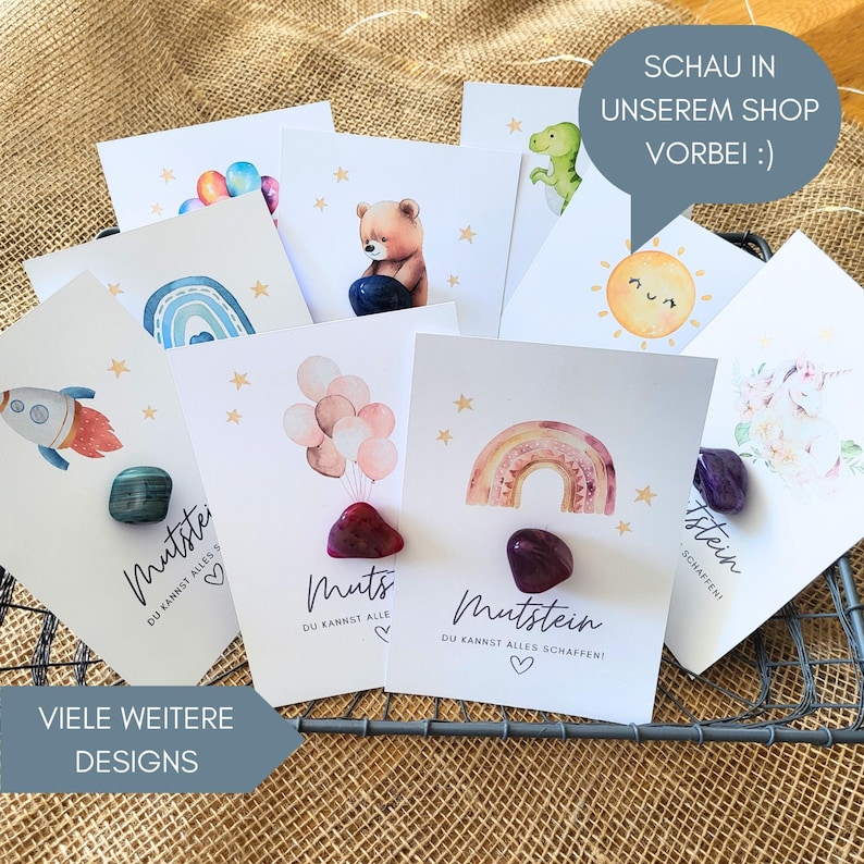 Op de afbeelding: Een verzameling witte kaarten met aquarelillustraties van raketten, regenbogen, ballonnen, beren en zonnen, elk gecombineerd met een gepolijste edelsteen. De kaarten tonen de Duitse tekst Mutstein en Du kannst alles schaffen.