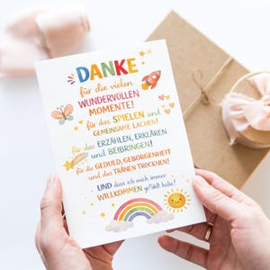 May include: A white greeting card with colorful text and illustrations, including a rocket, butterfly, hearts, and a rainbow. The card says "DANKE" and other German text. The card is held in front of a wrapped gift.