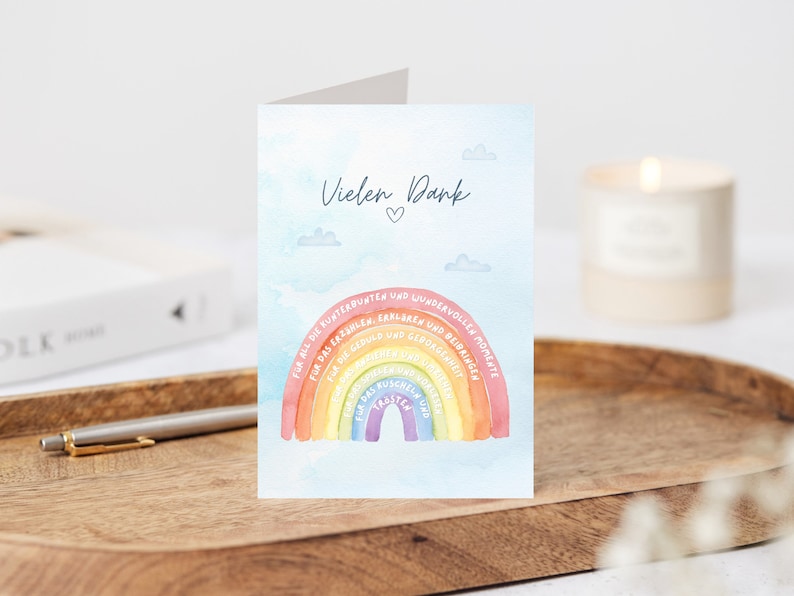 Op de afbeelding: Een kaart met een regenboogontwerp en de tekst "Vielen Dank" (Duits voor "Bedankt") met een hartje. De regenboog heeft tekst op elke band, waaronder "Voor al het plezier", "Voor het lachen", "Voor de herinneringen", "Voor het geduld", "Voor de liefde", "Voor de steun", "Voor de vriendelijkheid", "Voor de knuffels", "Voor de glimlachen", "Voor de vreugde", "Voor de magie", "Voor het wonder", "Voor de momenten".
