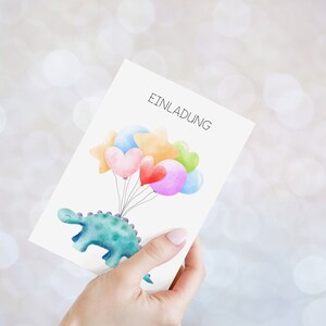 May include: A white card with the word "Einladung" printed on it. A green dinosaur with a blue belly is holding a string of colorful balloons, including hearts and stars.