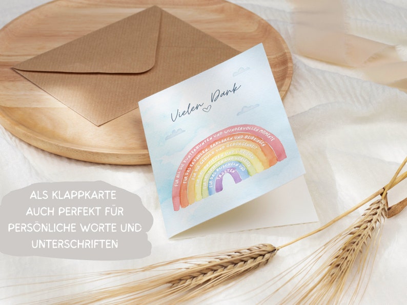 Op de afbeelding: Een gevouwen kaart met een aquarel regenboogontwerp en de tekst "Vielen Dank" (Duits voor "Bedankt"). De regenboog heeft Duitse woorden erop geschreven. De kaart ligt op een houten oppervlak met een bruine envelop en tarwehalmen.