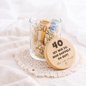 May include: A clear glass jar filled with dried white flowers and rolled-up Euro banknotes. A round wooden lid rests in front, engraved with the text 40 IST WIE 20, NUR DOPPELT SO GUT! ALLES LIEBE MIRELA and a small heart symbol.