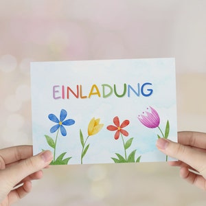 May include: A white card with the word "EINLADUNG" written in rainbow colors. The card is decorated with colorful flowers and has a watercolor background.