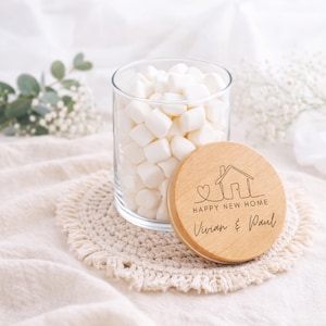 Könnte beinhalten: Ein klares Glas mit weißen Marshmallows und einem Holzdeckel. Der Deckel ist mit einer Hausgrafik, den Worten "HAPPY NEW HOME" und den Namen "Vivian & Paul" graviert. Das Glas steht auf einem beigefarbenen Häkeluntersetzer.