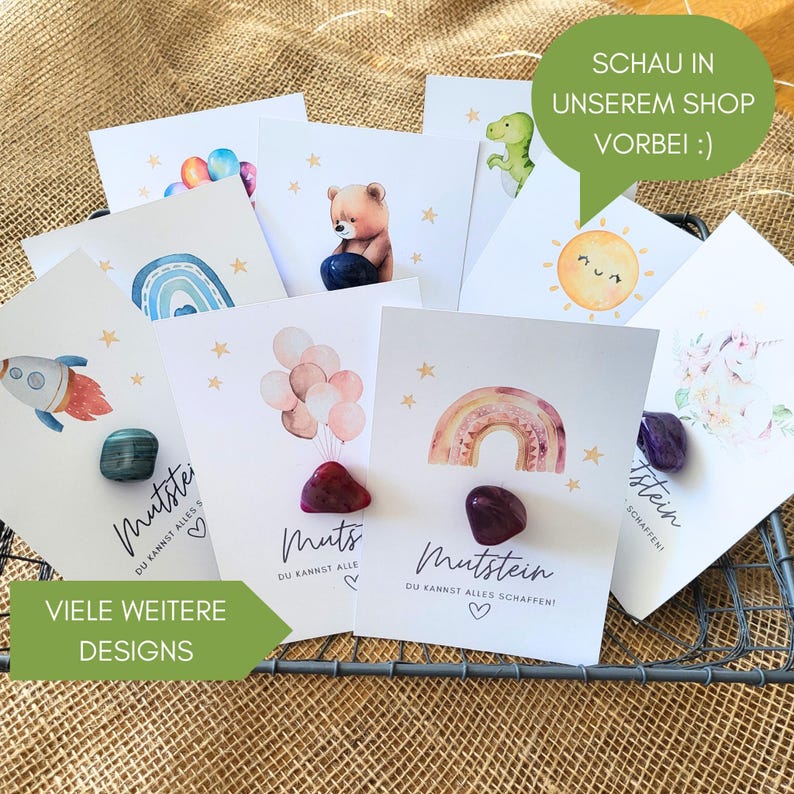 May include: A collection of white cards featuring watercolor illustrations of rockets, rainbows, balloons, bears, and suns, each paired with a polished gemstone and the German text Mutstein, Du kannst alles schaffen.