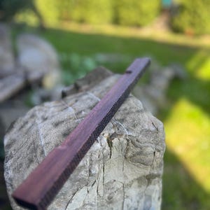 Puede incluir: Un largo taco de madera marrón oscuro con una superficie texturizada. El taco está apoyado en una roca gris.