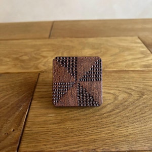 Puede incluir: Un cuadrado de madera marrón oscuro con un patrón geométrico tallado en la superficie. El patrón está formado por pequeños círculos y triángulos.