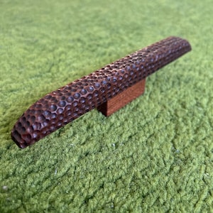 May include: A wooden guitar bridge with a textured surface. The bridge is brown and has a small rectangular base.