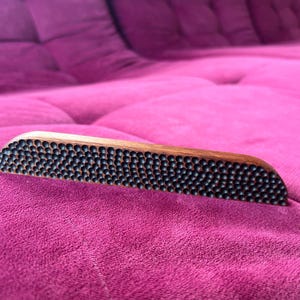 Puede incluir: Una herramienta de masaje de madera con una superficie texturizada de pequeños puntos negros. La herramienta está apoyada sobre una superficie de tela rosa.