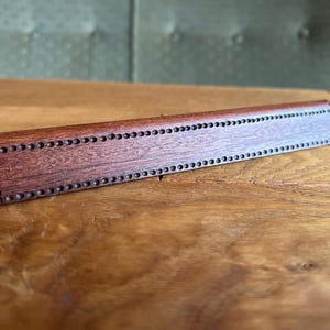 Puede incluir: Una placa de nombre de madera con un borde decorativo de pequeños círculos espaciados uniformemente. La placa de nombre es de un color marrón rojizo intenso.