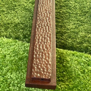 Puede incluir: Un acero de afilar rectangular de madera con una superficie texturizada. El acero es marrón y tiene un tono ligeramente rojizo.