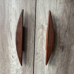 Puede incluir: Dos tiradores de armario de madera con un diseño curvo en forma de hoja. Los tiradores son marrones con un ribete marrón oscuro. El tirador de la izquierda está etiquetado como "variante A" y el tirador de la derecha está etiquetado como "variante B".