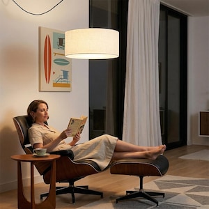 May include: A modern living room scene featuring a woman reading in a black leather chair with a matching ottoman. A round, beige lampshade hangs above. A small wooden side table holds a cup and saucer. A painting of surfboards and beach scenes hangs on the wall.
