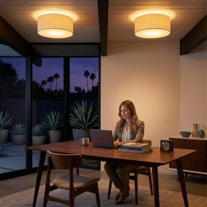 May include: Two round, beige fabric ceiling lights illuminate a home office. A woman sits at a dark wood table, working on a laptop. The room features a large window with a view of palm trees and potted plants.