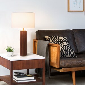 May include: A brown wooden table lamp with a white lampshade sits on a wooden end table with a white top. A small potted plant is on the table. A brown leather sofa with a black and white patterned pillow is in the background.