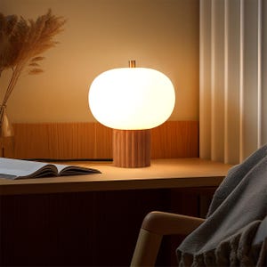 May include: A modern table lamp with a white, oval-shaped shade and a ribbed wooden base. The lamp is illuminated, casting a warm glow. The lamp sits on a wooden desk, next to an open book and a vase of dried flowers. The overall theme is home decor.
