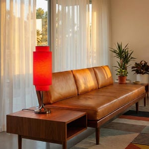 May include: A mid-century modern living room scene featuring a red cylindrical lamp on a walnut side table. A brown leather sofa with three cushions is positioned against a window with sheer curtains. Plants and a patterned rug add to the room's decor.