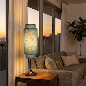 May include: A teal blue, cylindrical floor lamp with a textured shade, set on a dark metal base. The lamp is on a wooden side table in a living room with a sofa and large windows. The room has a warm, inviting atmosphere.