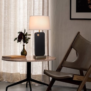 May include: A table lamp with a rectangular matte black base, a metallic silver square bottom, and a white fabric drum shade. The lamp features a small circular gold accent on the upper front of the black body and emits a soft warm light.