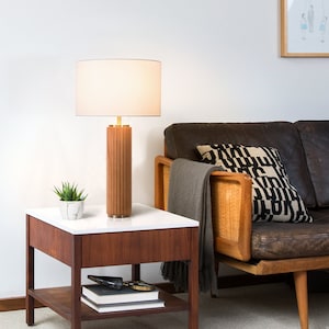 May include: A wooden table lamp with a white lampshade and a fluted wooden base. The lamp is on a wooden end table with a white top and a lower shelf. A small potted plant is on the table next to the lamp.