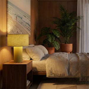 May include: A bedroom scene featuring a green table lamp with a cylindrical base and a rectangular lampshade. The lamp sits on a dark wooden nightstand next to a bed with neutral-colored bedding. Potted plants and a beach scene artwork complete the cozy interior.