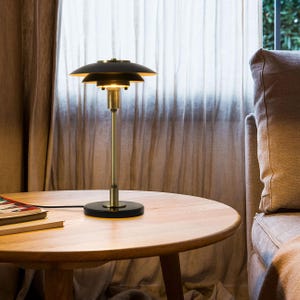 May include: A table lamp featuring a matte black multi-tiered shade, a brass-toned central stem, and a circular black base. The lamp emits a warm glow from beneath the shades, with a black power cord extending from the base across the surface.