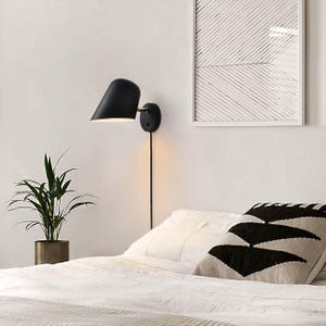 May include: A black wall-mounted sconce with a cone-shaped shade, mounted on a white wall. The sconce is connected to a black cord. A framed abstract art piece and a potted plant are also visible.
