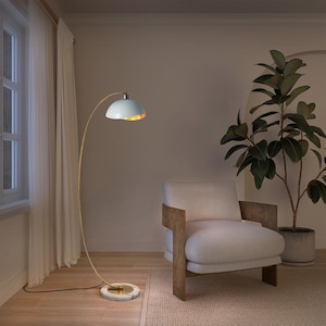 May include: A modern floor lamp with a white lampshade and a gold-colored, curved stem. The lamp has a marble base and is positioned near a white armchair and a potted plant. The room has a neutral color palette.