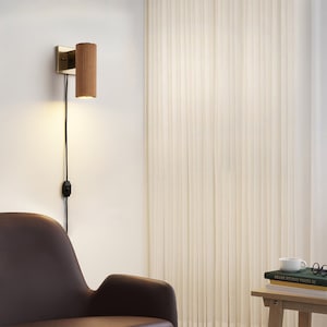 May include: A wall-mounted light fixture with a wooden shade and a black cord. The light is turned on and illuminating the room. The light is mounted on a white wall next to a white curtain.