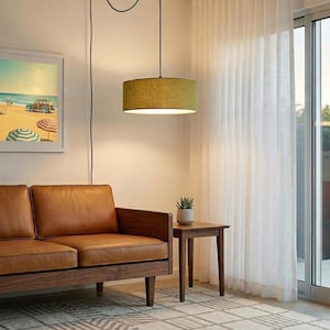 May include: A modern living room featuring a brown leather sofa, a small wooden side table, and a hanging green lampshade. A framed beach scene painting hangs on the wall. Sheer white curtains and a glass door are visible.