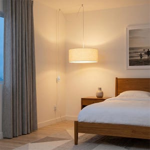 May include: A beige drum pendant light hangs from the ceiling in a bedroom. The light fixture is suspended by a white cord and is positioned above a wooden bed frame. A framed picture and a nightstand with a vase are also visible.