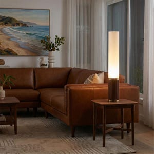 May include: A living room scene featuring a brown leather sectional sofa, a wooden side table, and a tall floor lamp. The lamp has a cylindrical design with a light-colored shade and a dark wood base. A painting of a beach scene hangs on the wall.