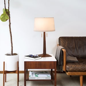May include: A white lamp with a brown wooden base and a white linen shade sits on a wooden end table with a book and glasses on top. A brown leather sofa with a throw blanket is in the background.