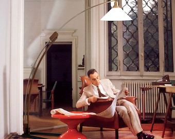May include: A mid-century modern arc floor lamp with a brass finish and a white, triangular shade. The lamp is positioned next to a red side table and a leather armchair. A person is seated in the chair reading a newspaper.