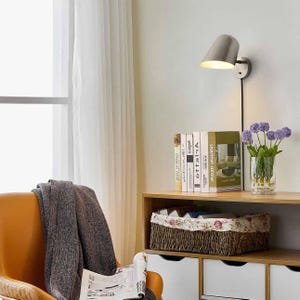 May include: A brushed metal wall-mounted lamp with a cone-shaped shade is mounted on a white wall. The lamp is positioned above a wooden shelf holding books, a floral basket, and a vase of purple flowers. A tan chair and a window with white curtains are in the background.