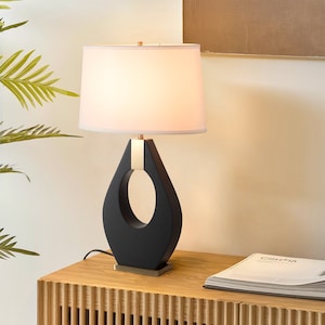 May include: A black table lamp with a white lampshade. The lamp has a teardrop-shaped base with a gold accent and a rectangular gold base. The lamp is on a wooden table.