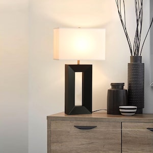 May include: A black rectangular table lamp with a white lampshade. The lamp is on a wooden cabinet with decorative items, including a black vase with a textured pattern, a small black jar, and a bowl with black stripes.