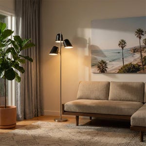 May include: A modern floor lamp with three black cone-shaped shades and a silver pole. The lamp is positioned in a living room with a beige sofa, a patterned rug, and a large potted plant. A coastal landscape painting hangs on the wall.